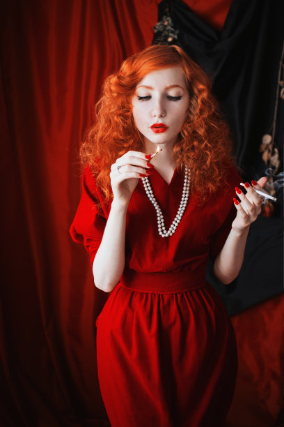一个红头发的女人穿着一件红色的合身的衣服，嘴里叼着一根香烟。红头发的女孩，苍白的皮肤和蓝色的眼睛，一个明亮的不寻常的外表，她的脖子上戴着珠子。黑色图片图片下载