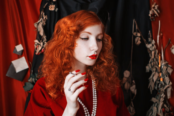 一个红头发的女人穿着一件红色的合身的衣服，嘴里叼着一根香烟。红头发的女孩，苍白的皮肤和蓝色的眼睛，一个明亮的不寻常的外表，她的脖子上戴着珠子。黑色图片图片下载