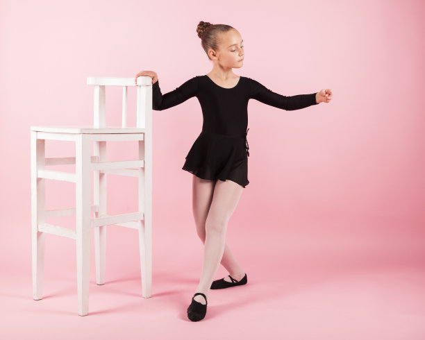 年轻美丽的女孩芭蕾舞女演员站在扶手椅附近练习芭蕾舞穿着黑色芭蕾舞裙摆在工作室与浅粉色的背景。图片下载
