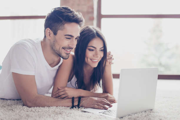 美丽的恋人在一起看蜜月的照片，拥抱着，躺在家里舒适的地毯上。他们很开心图片下载