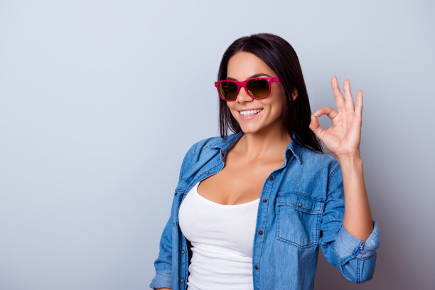 一个顽皮的年轻拉丁女孩的肖像在一个休闲的衣服和太阳镜，站在蓝色的背景和显示ok标志图片下载