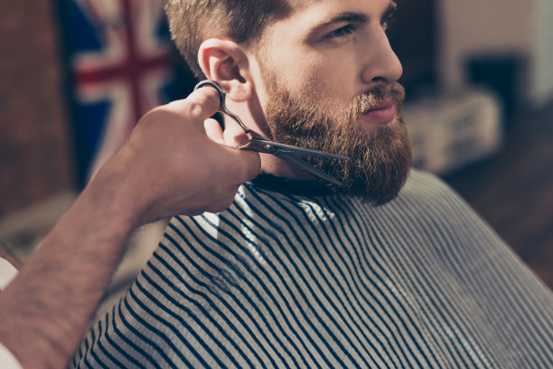 这是一个穿着条纹斗篷、留着红色大胡子的时髦男子在理发店里的特写照片，他的胡子被剪掉了，如此严肃和专注图片下载