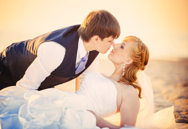 新婚夫妇躺在沙滩上，在夕阳下，一个穿着马甲的男人亲吻他的爱人，温暖和舒适的形象，休息和浪漫的关系图片下载