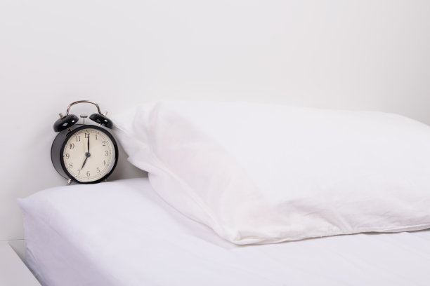 白色的床上放着一个黑色的闹钟，旁边是一个枕头图片下载