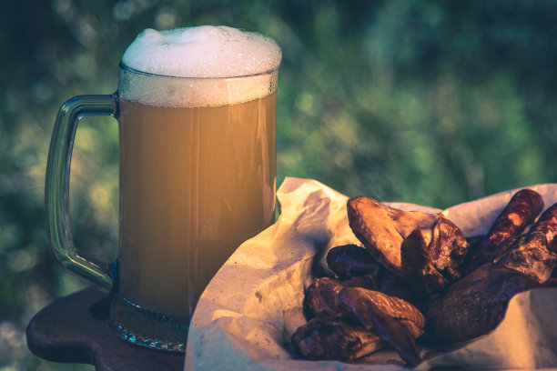 一杯淡啤酒，映衬着户外绿树。小吃香肠，熏鸡翅，复古色调图片图片下载