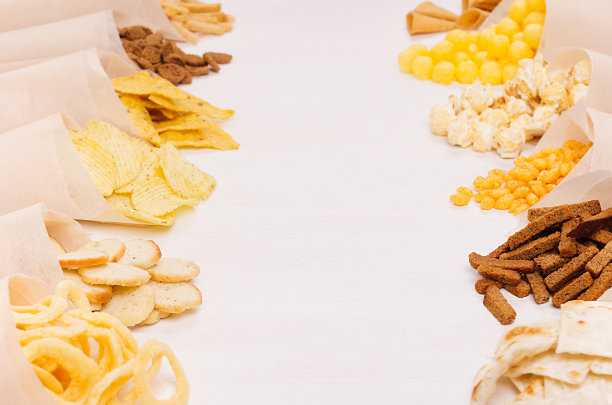 金色的彩色零食-玉米片，爆米花，油炸面包丁，薯片在手工纸锥作为边界的白色木桌与复制空间，特写，模糊。图片下载