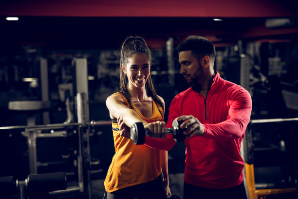 可爱的微笑肌肉健身女孩拿着一个哑铃在前面，做着肩部锻炼，看着镜头，而她帅气的私人教练站在她旁边，在健身房给她指导。图片下载
