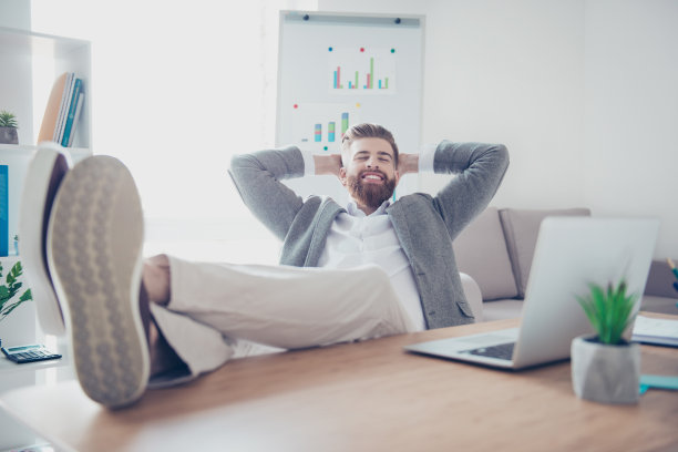 一个年轻开朗的商人正在工作场所休息，脚放在桌子上，面带微笑图片下载