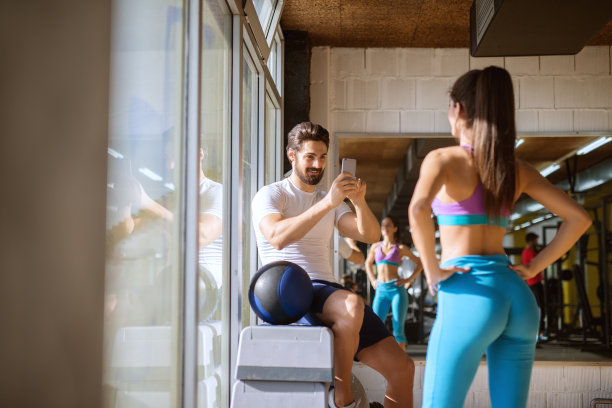 有魅力的蓄着胡须的英俊男子坐在健身房的角落里，站在靠近窗户的镜子前，用手机为他的运动健身女友拍照。图片下载