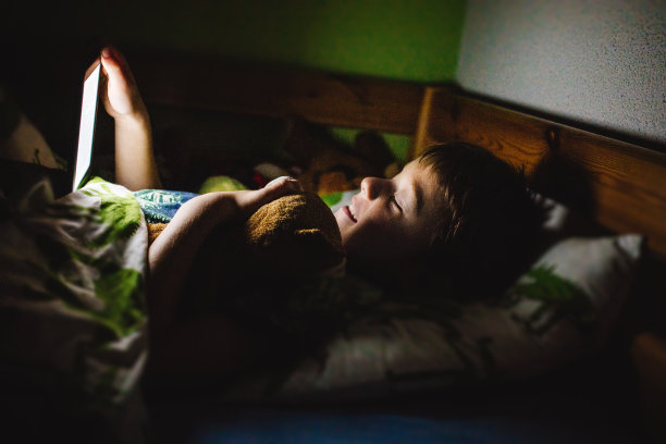 小男孩在睡觉前躺在床上看集成灯的电子书。儿童生活中的日常科技。背景暗，光线暗。图片下载