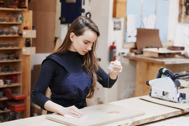 年轻快乐的妇女们站在木工车间的工作台上，给一棵树涂上清漆。家族企业。启动业务。年轻的专业设计师。图片下载