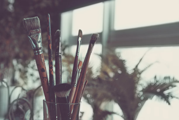 各种艺术家的画笔被放在一个透明的玻璃上，放在一个木制的乡村桌子上，桌子的背景是绿色的房间里的棕榈叶。在复古风格下加工。图片下载