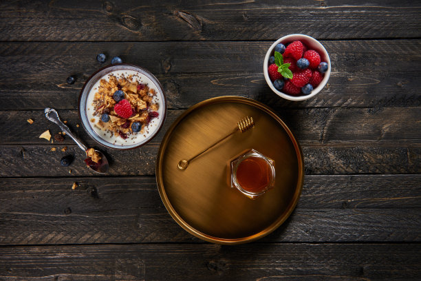 俯视图的健康早餐。牛奶什锦早餐，酸奶，蓝莓和覆盆子在碗和枫糖浆在金盘。深色木制背景。素材图片