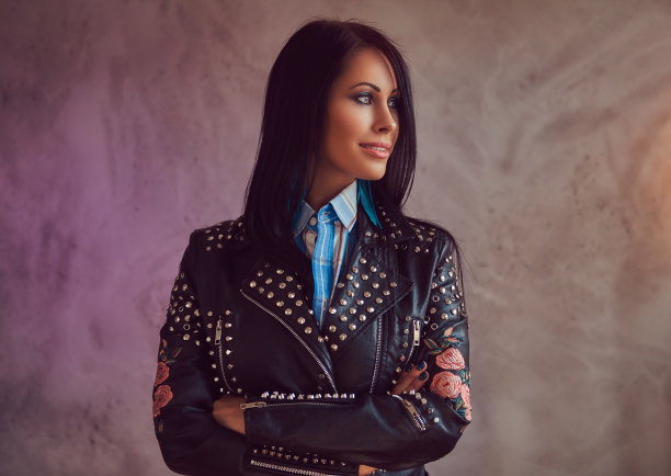 一个微笑的性感性感的黑发女子的肖像，穿着时尚的皮夹克和牛仔裤，双臂交叉在一个灰色的背景。图片下载