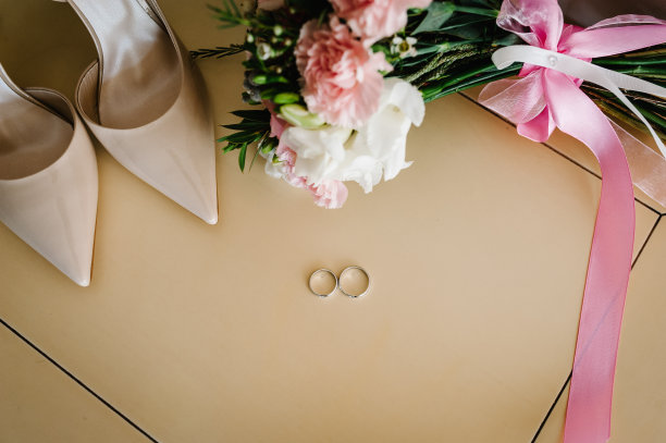 两个银色的结婚戒指，时尚优雅的经典漆米色鞋和婚礼花束的新娘，玫瑰和绿色躺在柔和的背景。关闭了。平的。前视图。图片下载