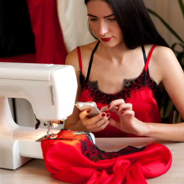 一位迷人的女裁缝穿着丝绸内衣坐在缝纫机前。缝纫博客照片她的工作与智能手机。女性在线时尚业务的概念。图片下载