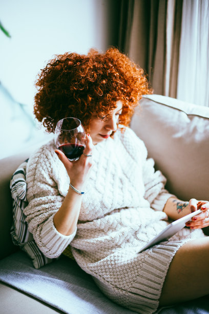 红发卷发的年轻女子，坐在沙发上，看着平板电脑屏幕，喝着红酒(调色图像，选择性聚焦)图片下载