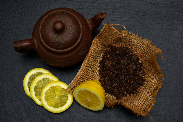 茶壶、半个和切片的成熟柠檬、黑色石板上的干燥茶叶、盘子或托盘素材图片