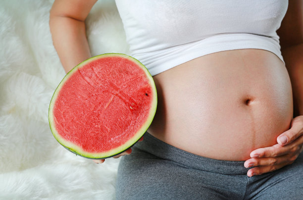 孕妇坐着，拿着切成一半的西瓜放在肚子上。图片下载