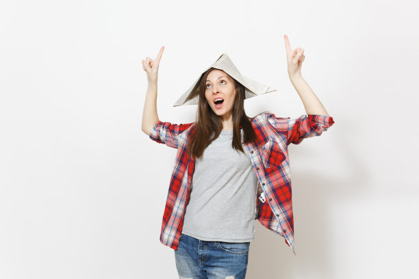 年轻的震惊美丽的女人在报纸帽子指向食指上的拷贝空间孤立的白色背景。公寓装修配件。修理家里的概念。广告区域。图片下载