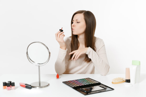 美丽的年轻女子用红色指甲油涂指甲，坐在桌子上使用脸部装饰化妆品隔离在白色的背景。美丽女性时尚生活理念。图片下载