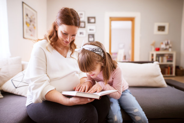 一个漂亮爱玩的女童坐在她细心的怀孕妈妈旁边，试图阅读这本书图片下载