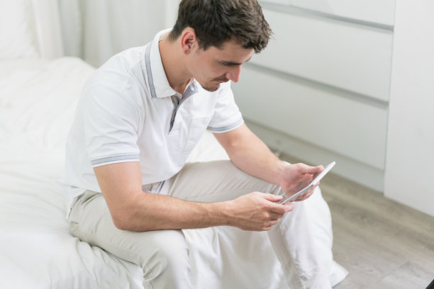 帅气的男人手里拿着平板电脑，坐在卧室的床上。男性在家里使用网络wifi。图片下载