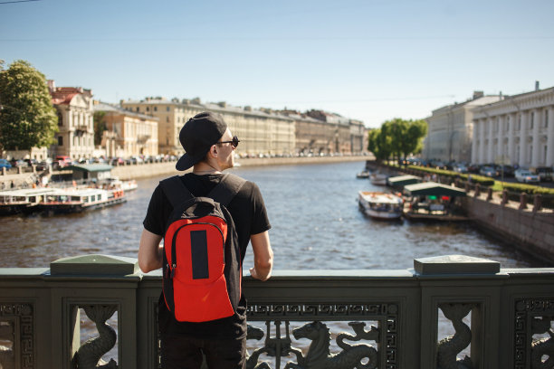 在俄罗斯圣彼得堡的格里博耶多夫运河堤岸上，时尚的男性游客背着背包欣赏城市风光图片下载