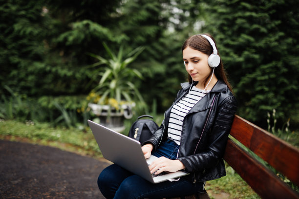 一张美丽的年轻女子坐在户外公园里的照片。用笔记本电脑戴着耳机听音乐。图片下载