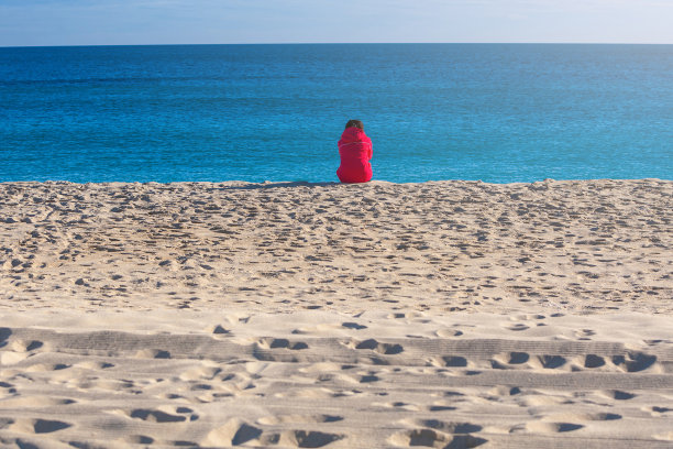 穿红夹克的女人坐在海滩上。晴朗的午后阳光，寒冷的天气里有沙滩和大海。图片下载
