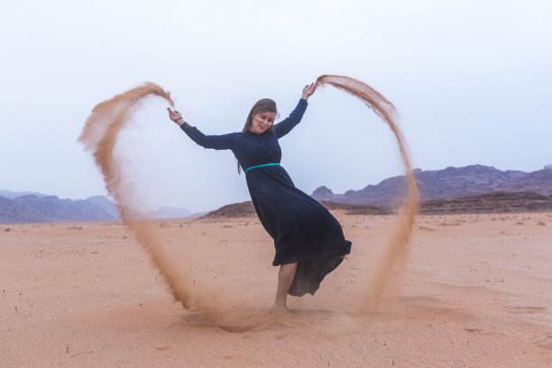 在约旦瓦迪拉姆沙漠，金发年轻女子身穿深蓝色长裙玩沙子图片下载