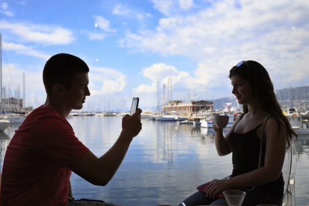 一对夫妇，年轻的男人和女人最好的朋友坐在咖啡馆，喝咖啡。用触摸屏，用智能手机拍照，照片短信。欣赏航海船、码头的景色。意大利的里雅斯特，亚得里亚海。图片下载