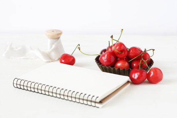 女性静物构图。甜美的红色樱桃水果在小金属碗，空白笔记本和一卷丝带在白色的桌子上。散焦背景。夏天的乡村场景。风格的库存图片。图片下载