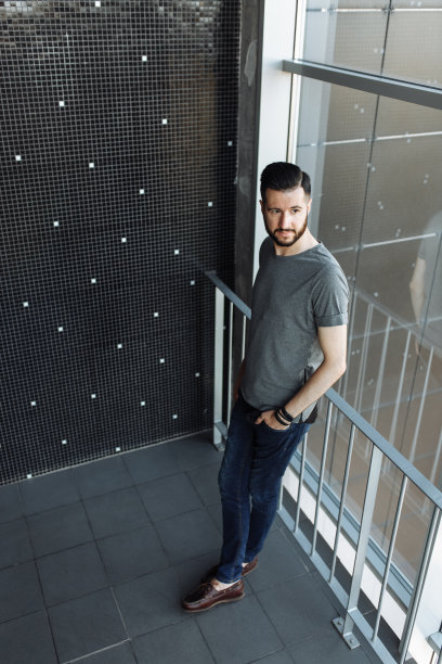 一个漂亮的时尚的男人的照片，潮人，穿着一件灰色空t恤，站在窗前，在一个黑色的墙壁背景。为标识或设计留出空间。图片下载