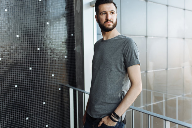 一个漂亮的时尚的男人的照片，潮人，穿着一件灰色空t恤，站在窗前，在一个黑色的墙壁背景。为标识或设计留出空间。图片下载