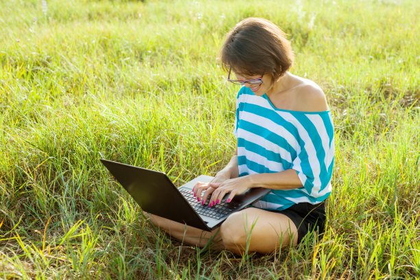 微笑的女孩坐在草地上在明亮的夏日和工作的电脑。在户外使用笔记本电脑，和朋友聊天。图片下载