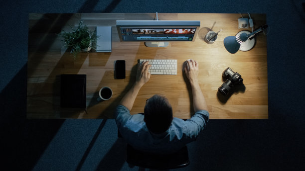 一个年轻的摄影师在深夜用他的台式电脑处理照片的俯视图。台灯照亮了他的桌子。他喝咖啡来保持清醒。相机、外置硬盘和笔记本电脑就在他身边。图片下载