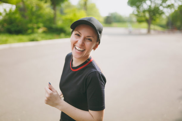 年轻的笑运动美丽的黑发女孩在黑色制服和帽子训练做运动，跑步和看在城市公园的道路上的摄像机。健身、健康的生活理念。图片下载