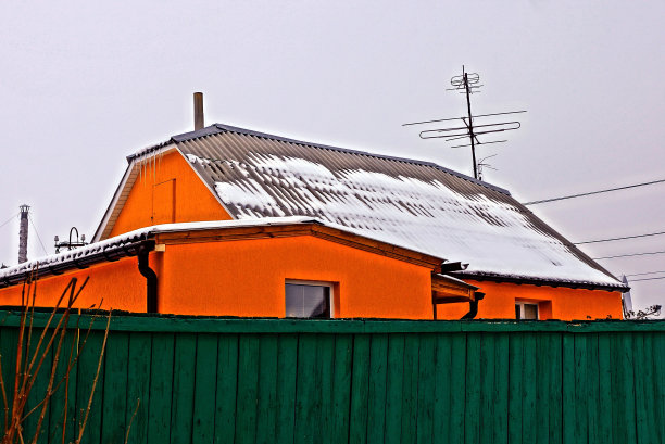 在绿色篱笆后面的雪地里橘黄色房子的一部分图片下载