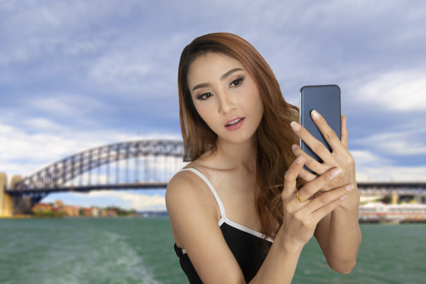 一名女性游客手持智能手机自拍，背景是桥和海。图片下载