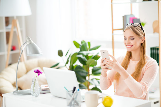 这是一幅欢快的女孩坐在桌前，在空闲时间拿着智能手机与朋友聊天，使用5G wi-fi网络进行视频通话的照片图片下载