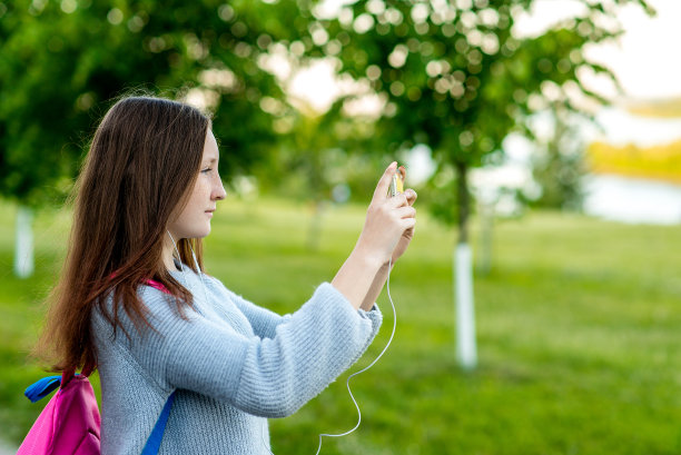 女孩女生少年。夏天在自然。他的背包后面拿着一部智能手机。照片在电话中。用耳机播放音乐。街头摄影的概念。图片下载