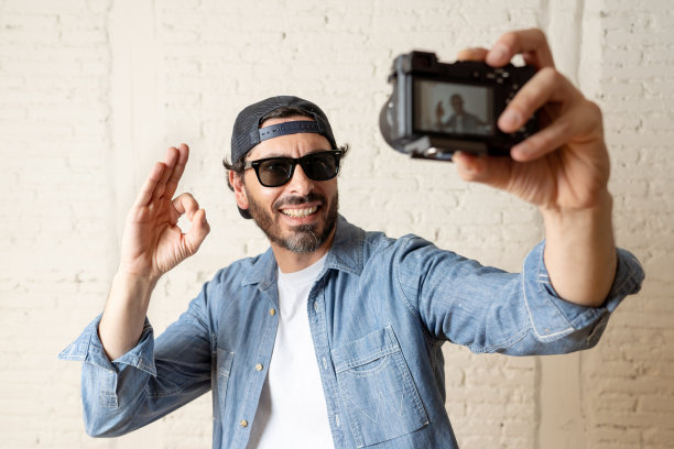 年轻有魅力的男人穿着休闲装，太阳镜在潮人风格的现代look拿着相机拍摄自画像自拍图片或在互联网博客和博客概念录制视频。图片下载