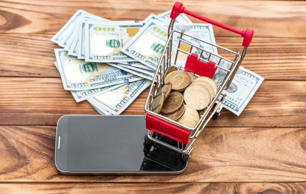 木桌上放着装有钱和智能手机的购物车。电子商务的概念。前视图。图片下载