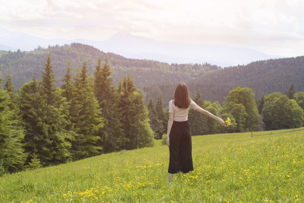 一位年轻女子手持一束鲜花站在草地上。森林和山脉的背景。后视图图片下载