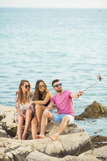 三个年轻的朋友坐在海边的岩石上自拍图片下载