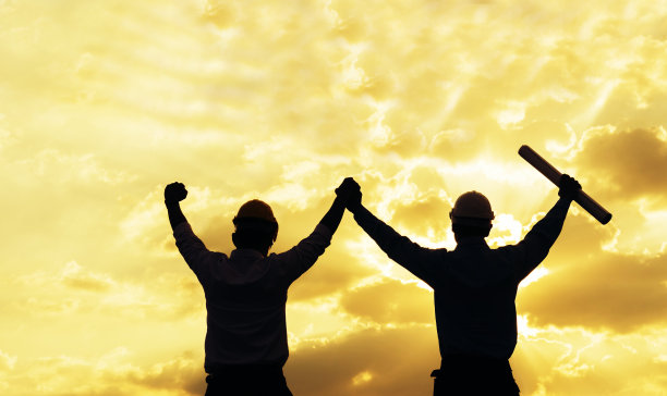 夕阳下成功工程师手牵手的剪影，胜利的标志。成功工程师戴上黄色安全帽，共同进行安全咨询。建筑的概念。图片下载