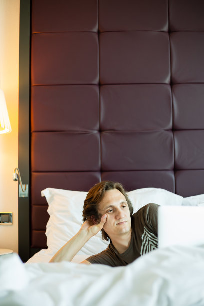 金发的年轻人躺在一张白色的床上，看着笔记本电脑的屏幕(选择性聚焦)图片下载