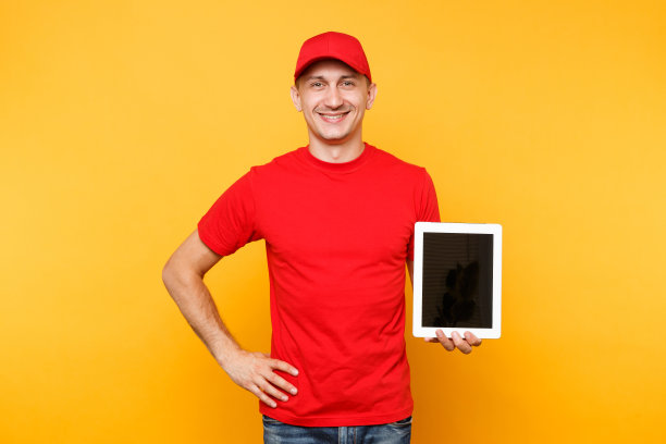 快递员被隔离在桔黄色背景上。职业男员工快递员，红帽，t恤，手持平板电脑与空白空白屏幕。服务的概念。副本的空间。模型模板。图片下载