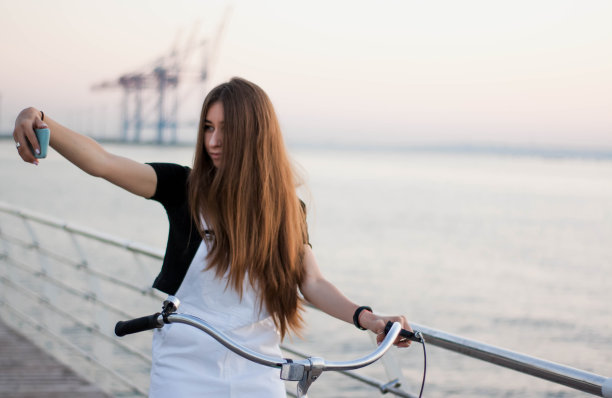 美丽的长发年轻女子骑着自行车玩得很开心，用智能手机，打电话，写短信，上网，在日落时自拍图片下载